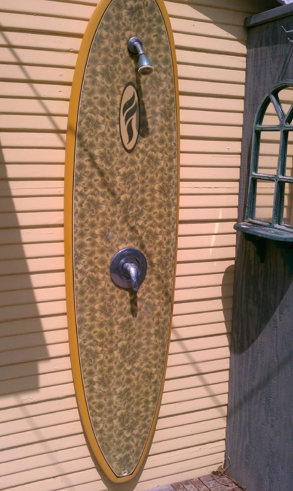 A surfboard converted into a shower, mounted on the exterior wall of a house, with water fixtures installed at the top and middle.
